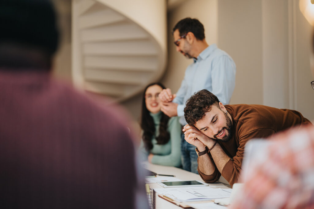 Mann sitzt am Tisch und hält Kopf in der Hand, andere Personen im Hintergrund in einem Raum mit Wendeltreppe.